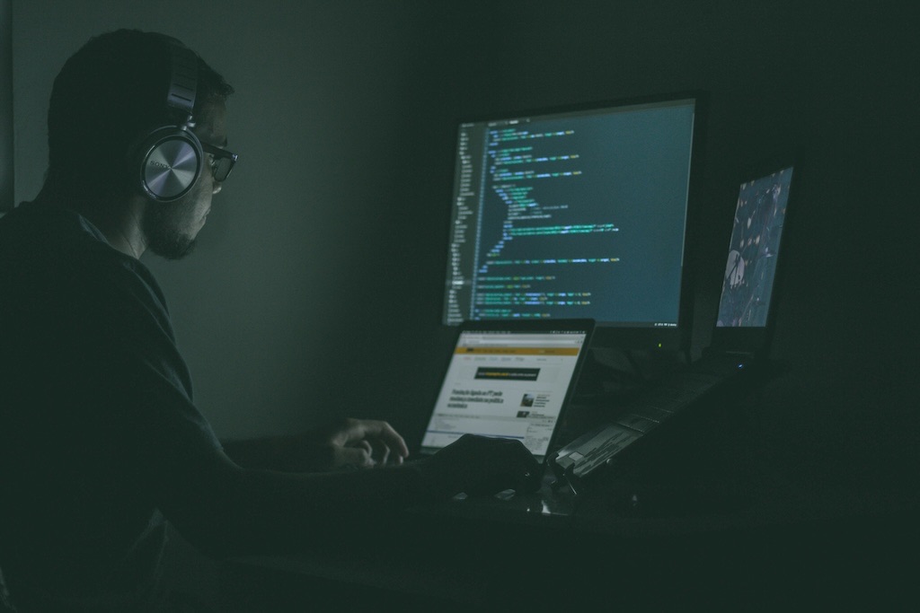 A developer in headphones working in a darkened room, lit only by the screens in front of them: a monitor of syntax-coloured code, an open laptop, and a third display showing a map
