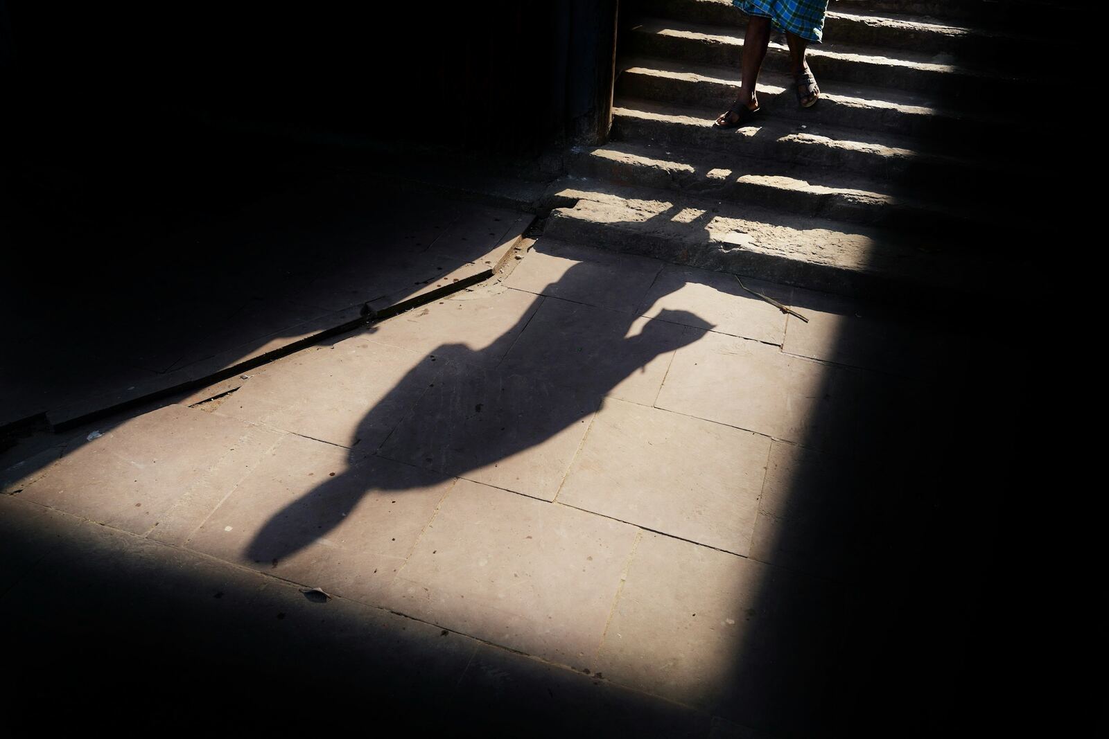 Hard sunlight across a stone floor, throwing the long shadow of a person whose own feet are visible on steps far behind it